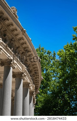 A dramatic low-angle view captures the sweeping curved entablature and ornate Corinthian capitals of a neoclassical Madrid colonnade. The elaborately carved stone cornice with sculptural figures curves against a deep blue sky while lush green summer trees fill the right side of the frame.