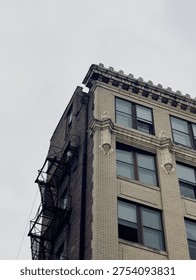 A dramatic low-angle view captures the weathered facade of an early 20th-century brick building in an urban setting, its beige brickwork contrasting with dark metal fire escapes clinging to the sides like industrial veins. Ornate cornices and decorative stone garlands crown the top, framing tall, narrow windows with subtle reflections of an overcast sky, evoking a sense of timeless resilience amid city decay. Soft, diffused gray light bathes the textured surfaces, highlighting peeling paint, rust stains, and intricate architectural details that whisper of bygone eras. The moody atmosphere blends nostalgia and grit, ideal for urban storytelling, architectural heritage illustrations, or atmospheric backdrops in editorial features on city evolution.