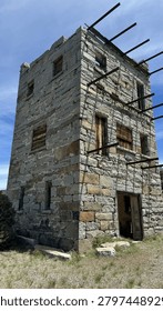 A dramatic low-angle view captures the well-preserved three-story granite stone tower of Stokes Castle standing against a vivid blue sky near Austin, Nevada. The historic 1897 Roman-style tower features weathered wooden window frames, rusted iron beam remnants, and richly textured stonework bathed in bright daylight. Ideal for travel editorial, American West history, ghost town, and Highway 50 road trip content.