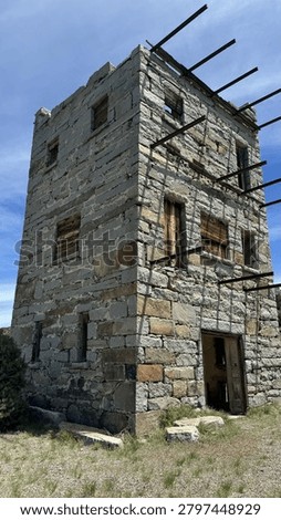 A dramatic low-angle view captures the well-preserved three-story granite stone tower of Stokes Castle standing against a vivid blue sky near Austin, Nevada. The historic 1897 Roman-style tower features weathered wooden window frames, rusted iron beam remnants, and richly textured stonework bathed in bright daylight. Ideal for travel editorial, American West history, ghost town, and Highway 50 road trip content.