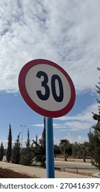 Dramatic low-angle view of a circular 30 speed limit sign on a blue post, contrasting sharply against a bright, cloud-filled blue sky above a paved roadway.