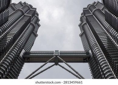 Dramatic low-angle view of the iconic Petronas Twin Towers and connecting skybridge under an overcast sky in Kuala Lumpur, Malaysia.