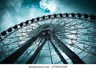 Dramatic low-angle view looking up at large Ferris wheel with gondola cabins against a moody teal dark cloudy sky, geometric steel structure detail.
