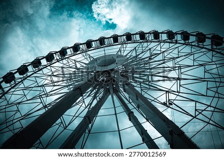 Dramatic low-angle view looking up at large Ferris wheel with gondola cabins against a moody teal dark cloudy sky, geometric steel structure detail.
