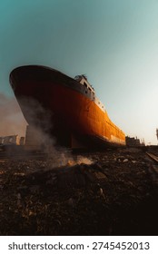 A dramatic low-angle view of a massive cargo ship resting at a shipbreaking yard during golden hour. Smoke rises from burning debris in the foreground, creating a powerful contrast against the warm sunlight illuminating the vessel’s rusted hull. Industrial buildings and urban structures appear in the background, symbolizing environmental impact, maritime industry, and urban decay.