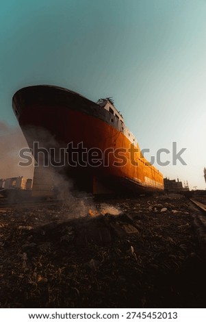 A dramatic low-angle view of a massive cargo ship resting at a shipbreaking yard during golden hour. Smoke rises from burning debris in the foreground, creating a powerful contrast against the warm sunlight illuminating the vessel’s rusted hull. Industrial buildings and urban structures appear in the background, symbolizing environmental impact, maritime industry, and urban decay.