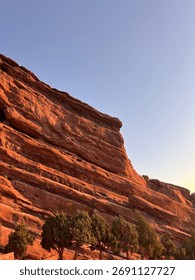 Dramatic low-angle view of massive red sandstone rock formations bathed in the golden light of sunrise or sunset, contrasting sharply with a clear blue sky.