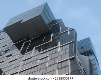 A dramatic low-angle view of a modern glass skyscraper featuring bold angular cantilevered sections and geometric architectural design against a clear blue sky.