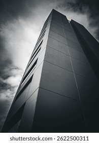 A dramatic, low-angle view of a modern high-rise building against a cloudy sky. The monochrome effect emphasizes the structure’s sharp lines and geometric patterns, a moody, architectural atmosphere.