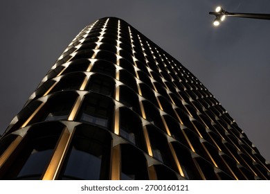 Dramatic low-angle view of a modern skyscraper at night. Its illuminated, curved facade creates a striking abstract pattern against a dark sky with copy space.