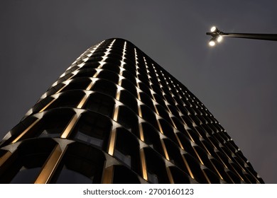Dramatic low-angle view of a modern skyscraper at night. Its illuminated, curved facade creates a striking abstract pattern against a dark sky with copy space.