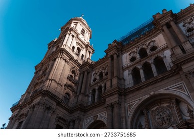 A dramatic low-angle view of the ornate Renaissance facade and the singular soaring tower of Malaga Cathedral against a deep blue sky in Spain.