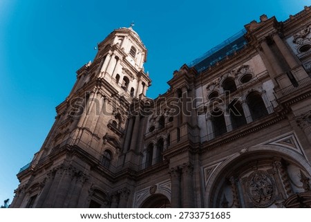 A dramatic low-angle view of the ornate Renaissance facade and the singular soaring tower of Malaga Cathedral against a deep blue sky in Spain.