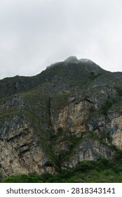 A dramatic low-angle view showcases a massive rocky mountain cliff face covered in sparse green vegetation under a cloudy sky, with wisps of fog clinging to the summit. The sheer stone texture, rugged crags, and deep crevices reflect nature’s raw power and geological beauty. Lush green foliage lines the bottom and scattered patches along the slope, offering a strong natural contrast against the dark gray rock. This vertical landscape image is ideal for adventure travel, nature documentaries, trekking, environmental topics, and outdoor exploration visual projects.