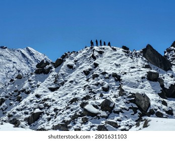 A dramatic low-angle view of a snow-covered rocky mountain ridge with a group of hikers standing silhouetted on the summit under a clear blue sky. The scene captures adventure, exploration, and the beauty of high-altitude winter landscapes, ideal for travel, trekking, and nature-themed projects.