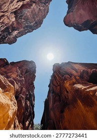 Dramatic low-angle view of towering red sandstone canyon walls under a bright sun and clear blue sky. Natural rock formations create a striking frame, highlighting geology, texture, and desert landscape beauty.