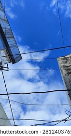 A dramatic low-angle, worm's-eye view composition frames a striking patch of vivid blue sky filled with fluffy white cumulus clouds. The natural beauty above contrasts sharply with concrete urban elements, featuring the textured concrete walls and sharp metallic awnings of city buildings creeping in from the edges. A complex network of black electrical power lines stretches horizontally across the frame, slicing through the view to create a gritty, modern city atmosphere. This street-level perspective is highly effective for editorial pieces discussing urban development, utility infrastructure, city living, or concepts exploring a literal 
