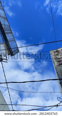 A dramatic low-angle, worm's-eye view composition frames a striking patch of vivid blue sky filled with fluffy white cumulus clouds. The natural beauty above contrasts sharply with concrete urban elements, featuring the textured concrete walls and sharp metallic awnings of city buildings creeping in from the edges. A complex network of black electrical power lines stretches horizontally across the frame, slicing through the view to create a gritty, modern city atmosphere. This street-level perspective is highly effective for editorial pieces discussing urban development, utility infrastructure, city living, or concepts exploring a literal 