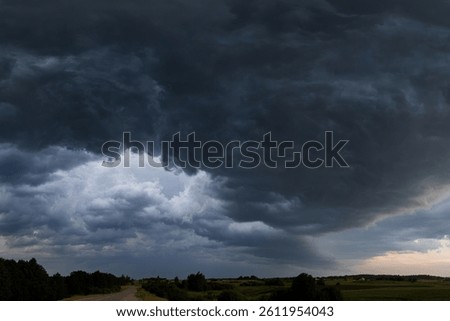 Dramatic mass of dark storm clouds descends over a quiet countryside road in Lithuania. The layered textures of the clouds convey the intensity of the approaching storm