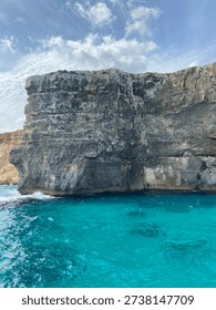 Dramatic massive limestone cliff face rising directly from vivid turquoise sea water. Layered rock formation with weathered surface texture photographed from a boat. White clouds in blue sky above. Clear Mediterranean water with waves breaking at the base of the cliff. Malta, Mediterranean Sea.