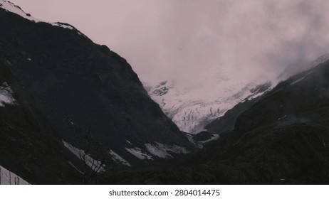 A dramatic and moody atmospheric shot featuring a towering, imposing mountain peak partially veiled in ethereal mist and low-hanging clouds. The striking contrast between the dark, rugged volcanic rock and the bright patches of snow creates a powerful, untamed wilderness vibe.