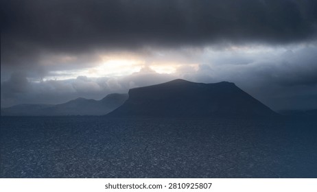 A dramatic and moody cloudscape featuring a dark silhouette of an island mountain under a heavy storm cloud sky with a golden breakthrough of sunlight on the horizon. Halloween background