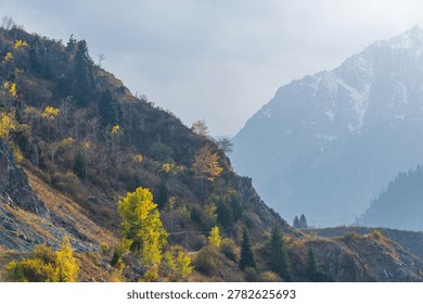 A dramatic, moody mountain scene featuring a sun-drenched, rocky foreground with autumn trees and hazy, snow-dusted peaks in the background valley.