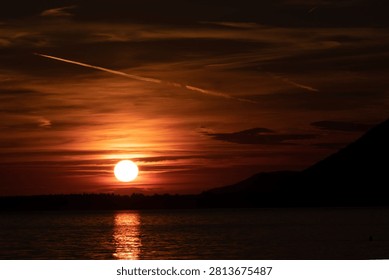 Dramatic moody sunset over the water with a bright sun, dark orange sky, and prominent mountain silhouette.
