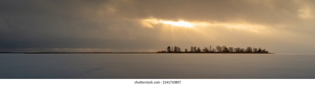 Dramatic Moody Winter Landscape. Wide Panoramic View Of A Frozen Snow-covered Lake With An Island Lit By Sunbeams Through A Gap Among A Cloudy Sky