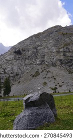 Dramatic mountain cliff face with evergreen trees and a large gray boulder on green grass, scale concept.