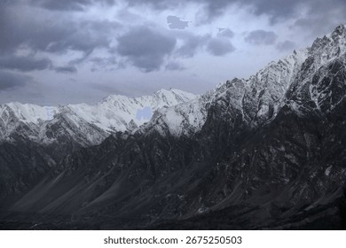 A dramatic mountain landscape features rugged peaks capped with snow, under a moody sky filled with clouds.