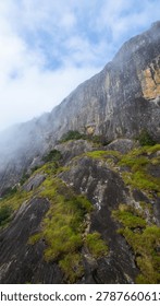 Dramatic mountain landscape featuring Pedra do Baú rising above dense fog and low clouds in the Serra da Mantiqueira region of southeastern Brazil. The massive rock formation dominates the scenery with steep walls and rugged terrain surrounded by forested hills and elevated peaks. Soft diffused light filters through mist covering the mountains, creating a cinematic atmosphere associated with adventure, exploration and remote highland environments.

The location is widely recognized for climbing, hiking and mountaineering activities, attracting visitors seeking outdoor experiences and panoramic views above the clouds. Trails crossing the mountainsides reveal changing weather conditions typical of high elevation regions, where fog and moving clouds constantly transform the scenery. The combination of dramatic relief, natural textures and expansive horizons creates a visually striking environment connected with ecotourism and adventure travel.

This scene was photographed at Pedra do Baú in the Serra da Mantiqueira, a famous destination in Brazil admired for rock climbing routes, mountain landscapes and scenic viewpoints surrounded by preserved nature in South America.