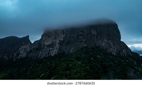 Dramatic mountain landscape featuring Pedra do Baú rising above dense fog and low clouds in the Serra da Mantiqueira region of southeastern Brazil. The massive rock formation dominates the scenery with steep walls and rugged terrain surrounded by forested hills and elevated peaks. Soft diffused light filters through mist covering the mountains, creating a cinematic atmosphere associated with adventure, exploration and remote highland environments.

The location is widely recognized for climbing, hiking and mountaineering activities, attracting visitors seeking outdoor experiences and panoramic views above the clouds. Trails crossing the mountainsides reveal changing weather conditions typical of high elevation regions, where fog and moving clouds constantly transform the scenery. The combination of dramatic relief, natural textures and expansive horizons creates a visually striking environment connected with ecotourism and adventure travel.

This scene was photographed at Pedra do Baú in the Serra da Mantiqueira, a famous destination in Brazil admired for rock climbing routes, mountain landscapes and scenic viewpoints surrounded by preserved nature in South America.