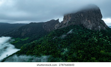 Dramatic mountain landscape featuring Pedra do Baú rising above dense fog and low clouds in the Serra da Mantiqueira region of southeastern Brazil. The massive rock formation dominates the scenery with steep walls and rugged terrain surrounded by forested hills and elevated peaks. Soft diffused light filters through mist covering the mountains, creating a cinematic atmosphere associated with adventure, exploration and remote highland environments.

The location is widely recognized for climbing, hiking and mountaineering activities, attracting visitors seeking outdoor experiences and panoramic views above the clouds. Trails crossing the mountainsides reveal changing weather conditions typical of high elevation regions, where fog and moving clouds constantly transform the scenery. The combination of dramatic relief, natural textures and expansive horizons creates a visually striking environment connected with ecotourism and adventure travel.

This scene was photographed at Pedra do Baú in the Serra da Mantiqueira, a famous destination in Brazil admired for rock climbing routes, mountain landscapes and scenic viewpoints surrounded by preserved nature in South America.