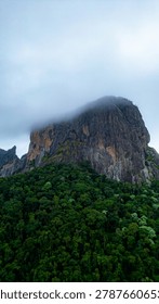 Dramatic mountain landscape featuring Pedra do Baú rising above dense fog and low clouds in the Serra da Mantiqueira region of southeastern Brazil. The massive rock formation dominates the scenery with steep walls and rugged terrain surrounded by forested hills and elevated peaks. Soft diffused light filters through mist covering the mountains, creating a cinematic atmosphere associated with adventure, exploration and remote highland environments.

The location is widely recognized for climbing, hiking and mountaineering activities, attracting visitors seeking outdoor experiences and panoramic views above the clouds. Trails crossing the mountainsides reveal changing weather conditions typical of high elevation regions, where fog and moving clouds constantly transform the scenery. The combination of dramatic relief, natural textures and expansive horizons creates a visually striking environment connected with ecotourism and adventure travel.

This scene was photographed at Pedra do Baú in the Serra da Mantiqueira, a famous destination in Brazil admired for rock climbing routes, mountain landscapes and scenic viewpoints surrounded by preserved nature in South America.