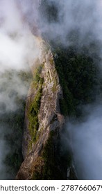 Dramatic mountain landscape featuring Pedra do Baú rising above dense fog and low clouds in the Serra da Mantiqueira region of southeastern Brazil. The massive rock formation dominates the scenery with steep walls and rugged terrain surrounded by forested hills and elevated peaks. Soft diffused light filters through mist covering the mountains, creating a cinematic atmosphere associated with adventure, exploration and remote highland environments.

The location is widely recognized for climbing, hiking and mountaineering activities, attracting visitors seeking outdoor experiences and panoramic views above the clouds. Trails crossing the mountainsides reveal changing weather conditions typical of high elevation regions, where fog and moving clouds constantly transform the scenery. The combination of dramatic relief, natural textures and expansive horizons creates a visually striking environment connected with ecotourism and adventure travel.

This scene was photographed at Pedra do Baú in the Serra da Mantiqueira, a famous destination in Brazil admired for rock climbing routes, mountain landscapes and scenic viewpoints surrounded by preserved nature in South America.