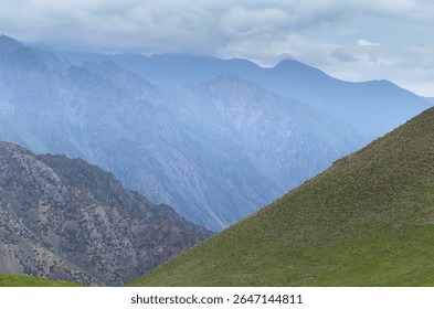 Dramatic mountain landscape featuring ranges receding into the distance with atmospheric haze.Foreground shows a green hillside contrasting with rugged rocky terrain and misty peaks under a cloudy sky