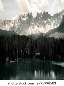 A dramatic mountain landscape featuring rugged snow-capped peaks towering above a tranquil alpine lake, surrounded by a dense evergreen forest. Soft clouds drift across the sky, creating a moody and atmospheric wilderness scene with reflections shimmering on the calm water.