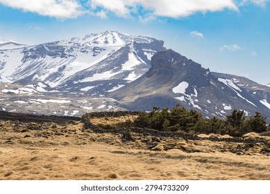 A dramatic mountain landscape featuring snow covered volcanic peaks and rugged terrain in Iceland. The scene showcases the raw geology and striking contrasts that make Iceland a popular travel destination.
