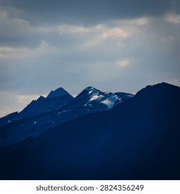 Dramatic mountain landscape featuring snow-capped peaks and deep blue silhouettes against a cloudy sky, evoking peace and solitude