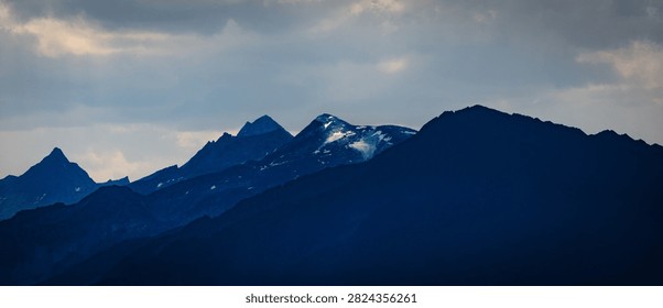 Dramatic mountain landscape featuring snow-capped peaks and deep blue silhouettes against a cloudy sky, evoking peace and solitude