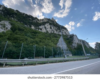 Dramatic mountain landscape featuring a steep, rocky cliff covered in lush green vegetation under a bright blue sky with scattered white clouds. In the foreground, an asphalt road with a metal guardrail runs alongside a fenced sports field with football goals. At the top of the mountain, a historic stone tower overlooks the valley. The scene captures the contrast between natural rugged terrain and human infrastructure in a scenic Italian countryside setting.