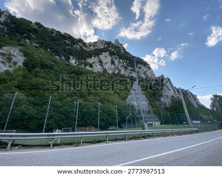 Dramatic mountain landscape featuring a steep, rocky cliff covered in lush green vegetation under a bright blue sky with scattered white clouds. In the foreground, an asphalt road with a metal guardrail runs alongside a fenced sports field with football goals. At the top of the mountain, a historic stone tower overlooks the valley. The scene captures the contrast between natural rugged terrain and human infrastructure in a scenic Italian countryside setting.
