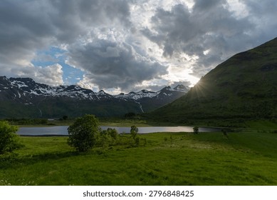 Dramatic mountain landscape featuring a tranquil lake, lush green meadows, and snow-capped peaks illuminated by the setting sun. Sun rays breaking through dense clouds create a breathtaking wilderness scene in a remote Nordic valley.