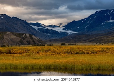 Dramatic mountain landscape with a glacier, fall foliage, and a reflective water surface under a cloudy sky.
