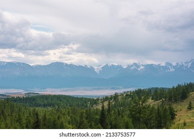 Dramatic mountain landscape with green coniferous forest hills and high snowy mountain range under rainy cloudy sky. Atmospheric aerial view to conifer forest and large snow mountains in overcast.