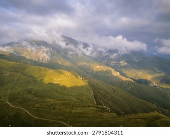 Dramatic mountain landscape with low clouds and golden light patches on green slopes, aerial view