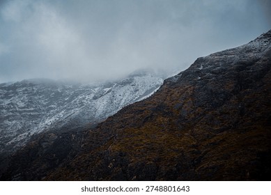 A dramatic mountain landscape showcasing a snow‑capped peak rising sharply against a moody blue sky. In the foreground, a rugged, golden‑brown hillside dotted with sparse vegetation leads to two tall electrical transmission towers with multiple power lines stretching across the scene. The contrast highlights the raw natural beauty of the highland terrain juxtaposed with industrial infrastructure.