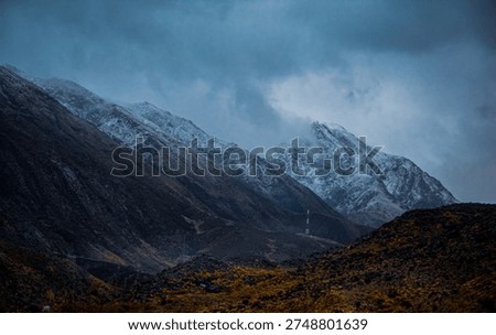 A dramatic mountain landscape showcasing a snow‑capped peak rising sharply against a moody blue sky. In the foreground, a rugged, golden‑brown hillside dotted with sparse vegetation leads to two tall electrical transmission towers with multiple power lines stretching across the scene. The contrast highlights the raw natural beauty of the highland terrain juxtaposed with industrial infrastructure.