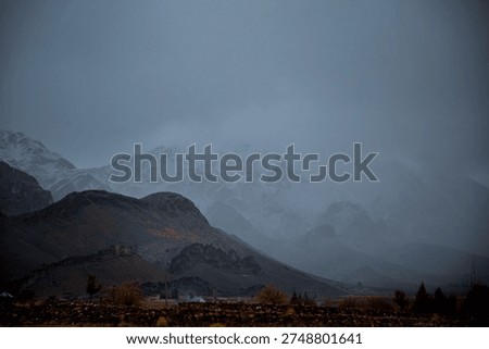 A dramatic mountain landscape showcasing a snow‑capped peak rising sharply against a moody blue sky. In the foreground, a rugged, golden‑brown hillside dotted with sparse vegetation leads to two tall electrical transmission towers with multiple power lines stretching across the scene. The contrast highlights the raw natural beauty of the highland terrain juxtaposed with industrial infrastructure.